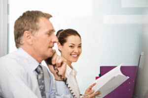 Two business professionals in an office, one man talking on the phone in focus foreground, and a smiling woman holding documents and a pen, representing teamwork, communication, and customer service support