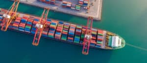 Aerial top view of a large container cargo ship docked at a port with colorful shipping containers and cranes loading and unloading goods on turquoise water