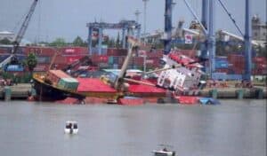 Contaminated cargo ship capsizes at a busy port with containers and cranes in the background, highlighting maritime logistics and shipping safety issues.