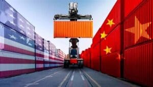 A container handler lifting an orange shipping container between stacks of cargo containers painted with the U.S. flag on the left and the Chinese flag on the right, symbolizing trade between the United States and China