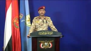Military spokesperson in camouflage uniform and red beret delivering a press conference at a podium with microphones, standing beside national and military flags