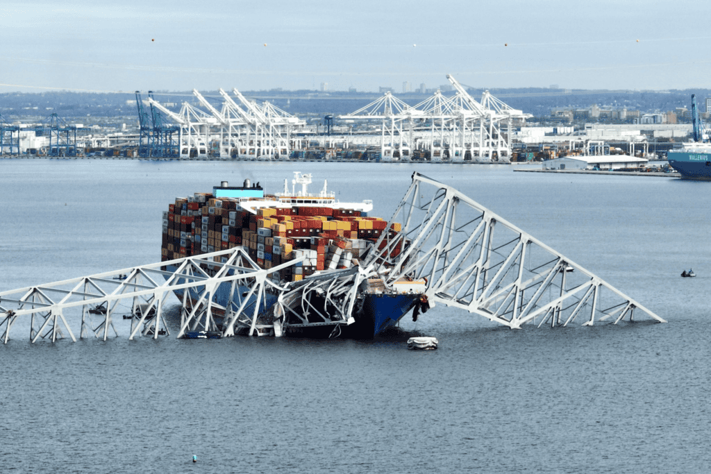 Container ship loaded with colorful shipping containers trapped under collapsed steel bridge structure in harbor with cranes in background