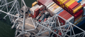 Close-up view of collapsed bridge structure resting on a container ship, with crushed shipping containers and twisted steel beams visible