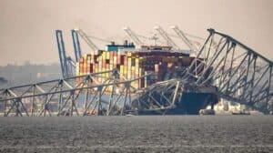 Container ship collision with collapsed steel bridge structure at a busy port, shipping containers stacked on deck visible in the background