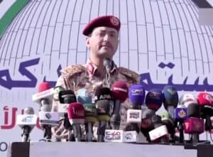 Military spokesperson in camouflage uniform addressing press conference with multiple microphones from various media outlets